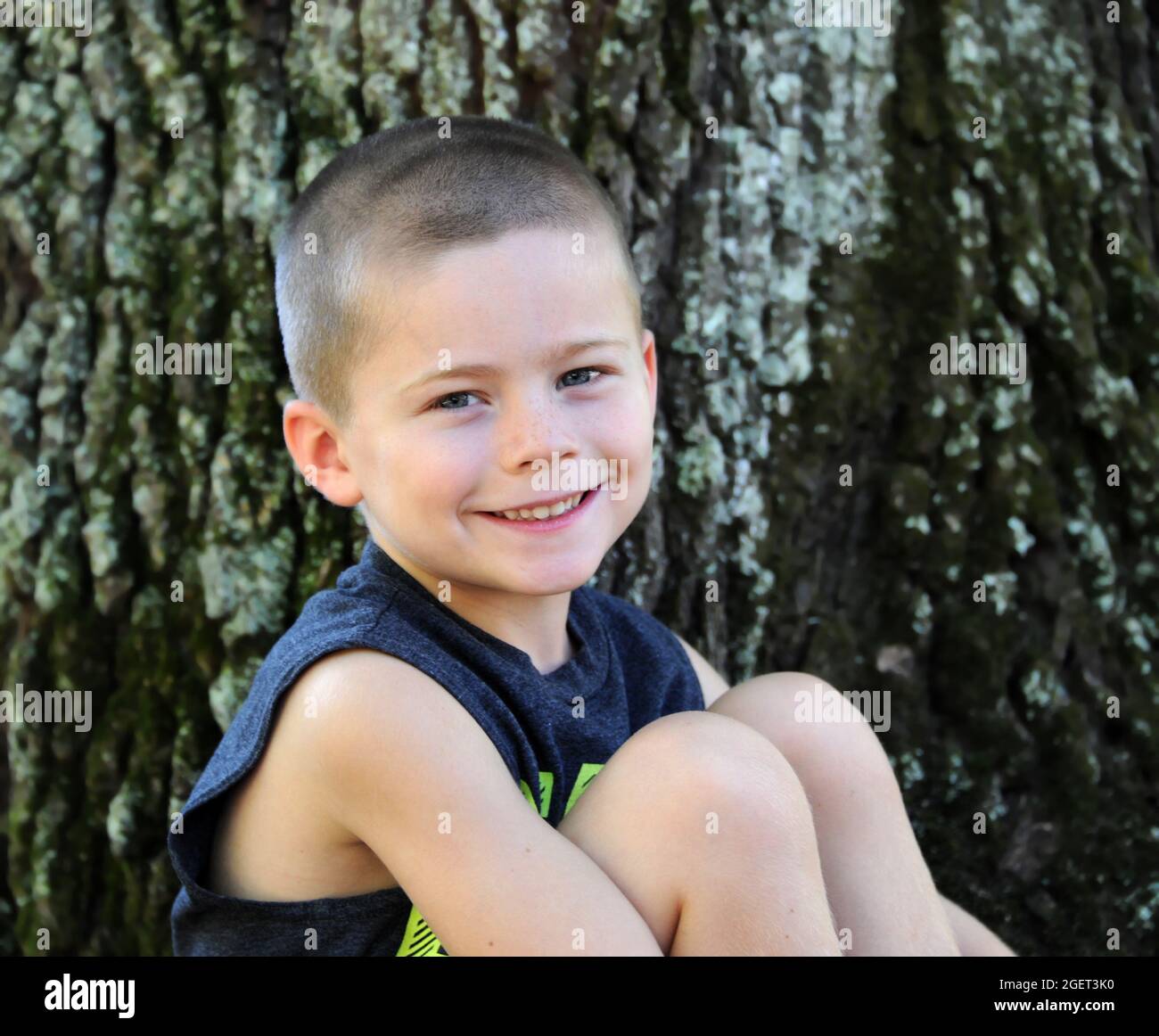 Il sorriso boyish si diffonde attraverso questo faccia dei ragazzi piccoli. È seduto al di fuori di un albero e guarda la macchina fotografica. Foto Stock
