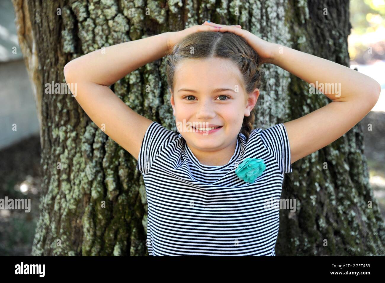 La bambina siede oltre un albero di quercia. Ha le mani sollevate dietro la testa e appoggiate contro il tronco dell'albero. Foto Stock