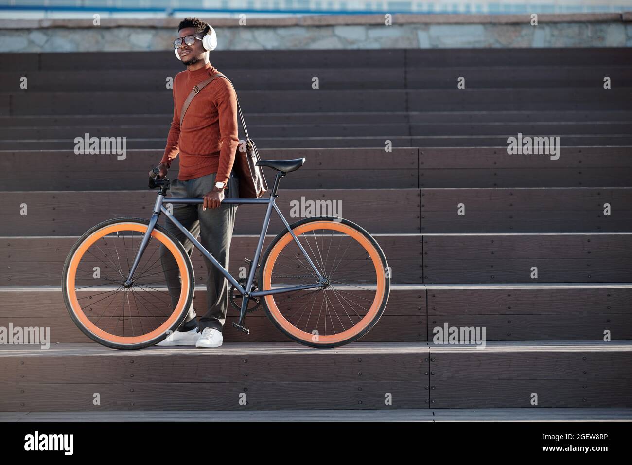 Giovane uomo d'affari in cuffie che ascolta musica mentre si alza sul gradino della scala con la sua bicicletta Foto Stock