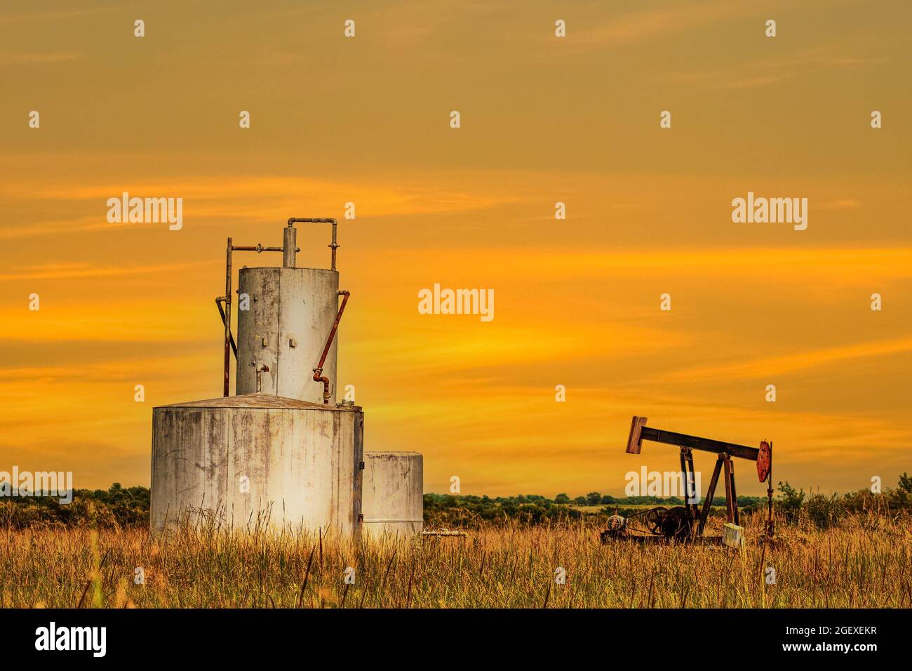 Carri armati e jack pompa in secco campo estivo con erbe arancioni che riflettono il cielo del tramonto con colline ondulate e alberi sullo sfondo. Foto Stock