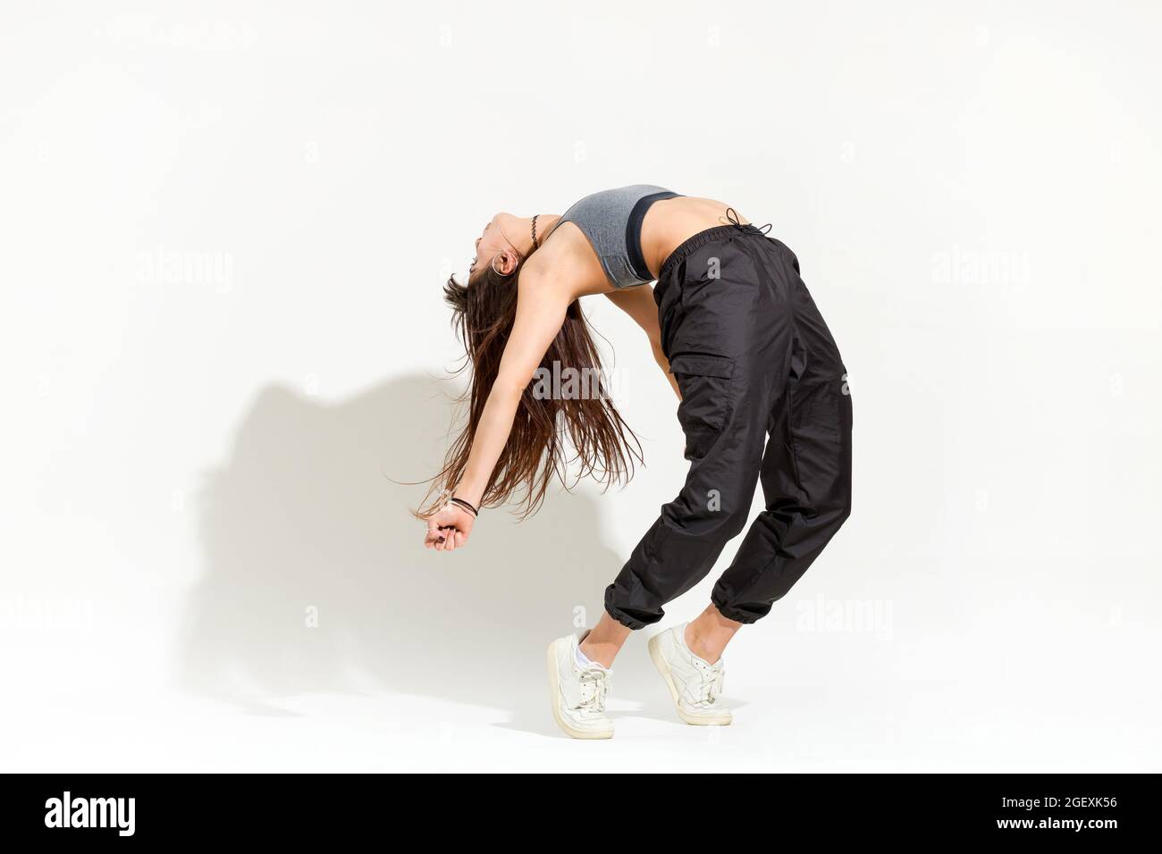 Agile giovane donna flessibile che fa una moderna danza hip hop posa equilibrare le sue dita dei piedi e arcate indietro gettando un'ombra su uno sfondo bianco Foto Stock