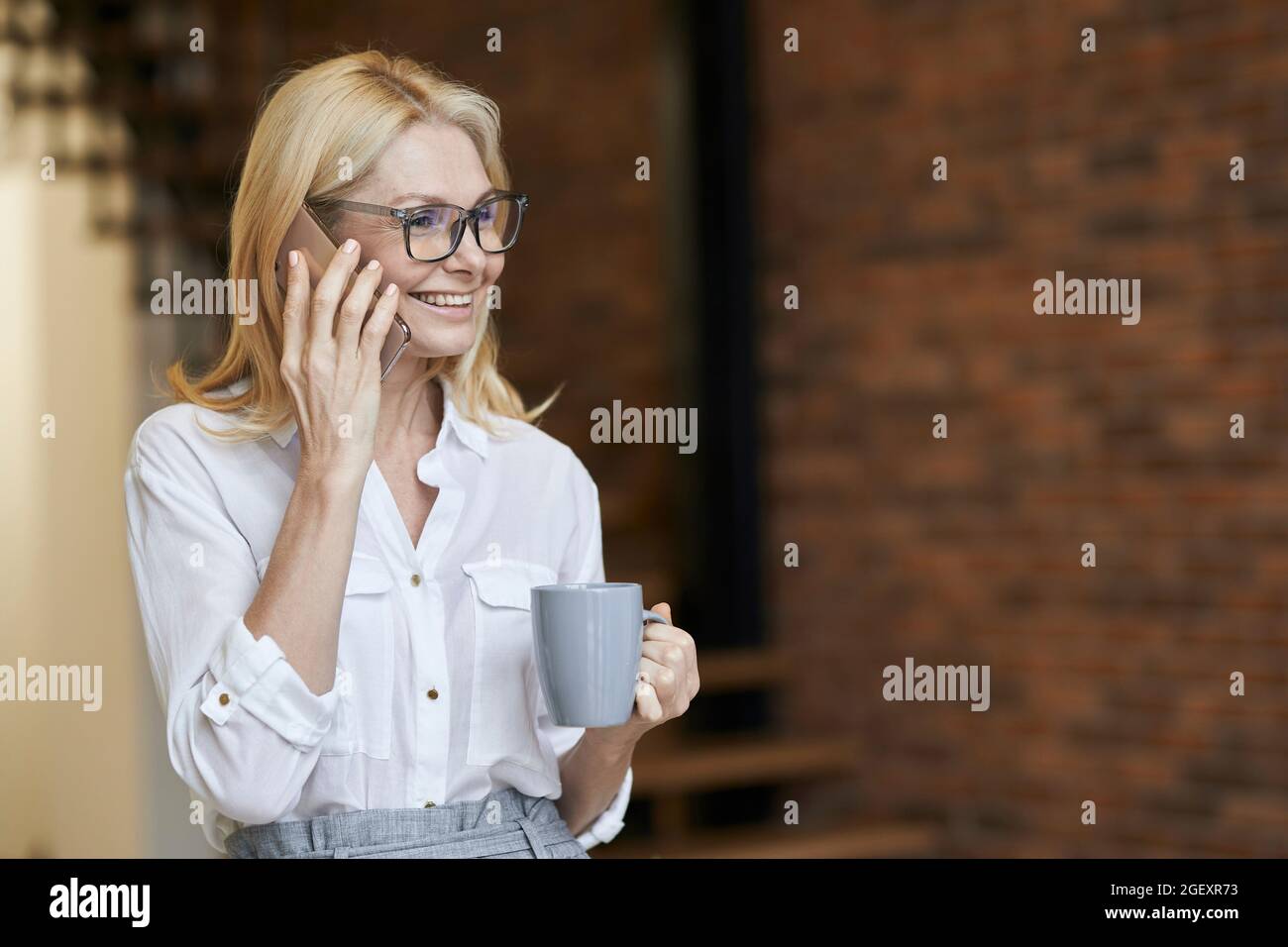 Donna occupata di mezza età con capelli biondi e bicchieri in camicia bianca bere caffè o tè mentre parla al telefono, in piedi a casa Foto Stock