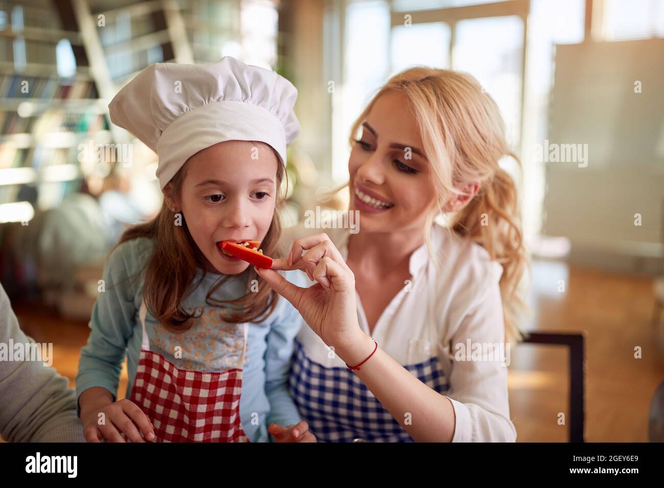 Una figlia piccola degusta le verdure date dalla madre mentre prepara un pasto in un'atmosfera allegra a casa insieme. Famiglia, tempo libero, insieme Foto Stock
