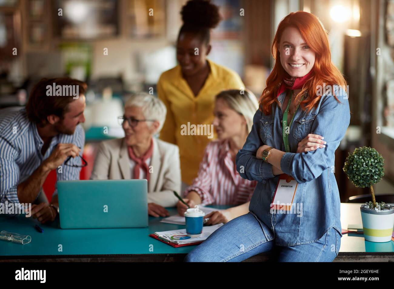 giovane donna rossa adulta che si pone al lavoro di fronte a un gruppo di lavoratori, sorridendo, guardando la macchina fotografica. contatto visivo Foto Stock