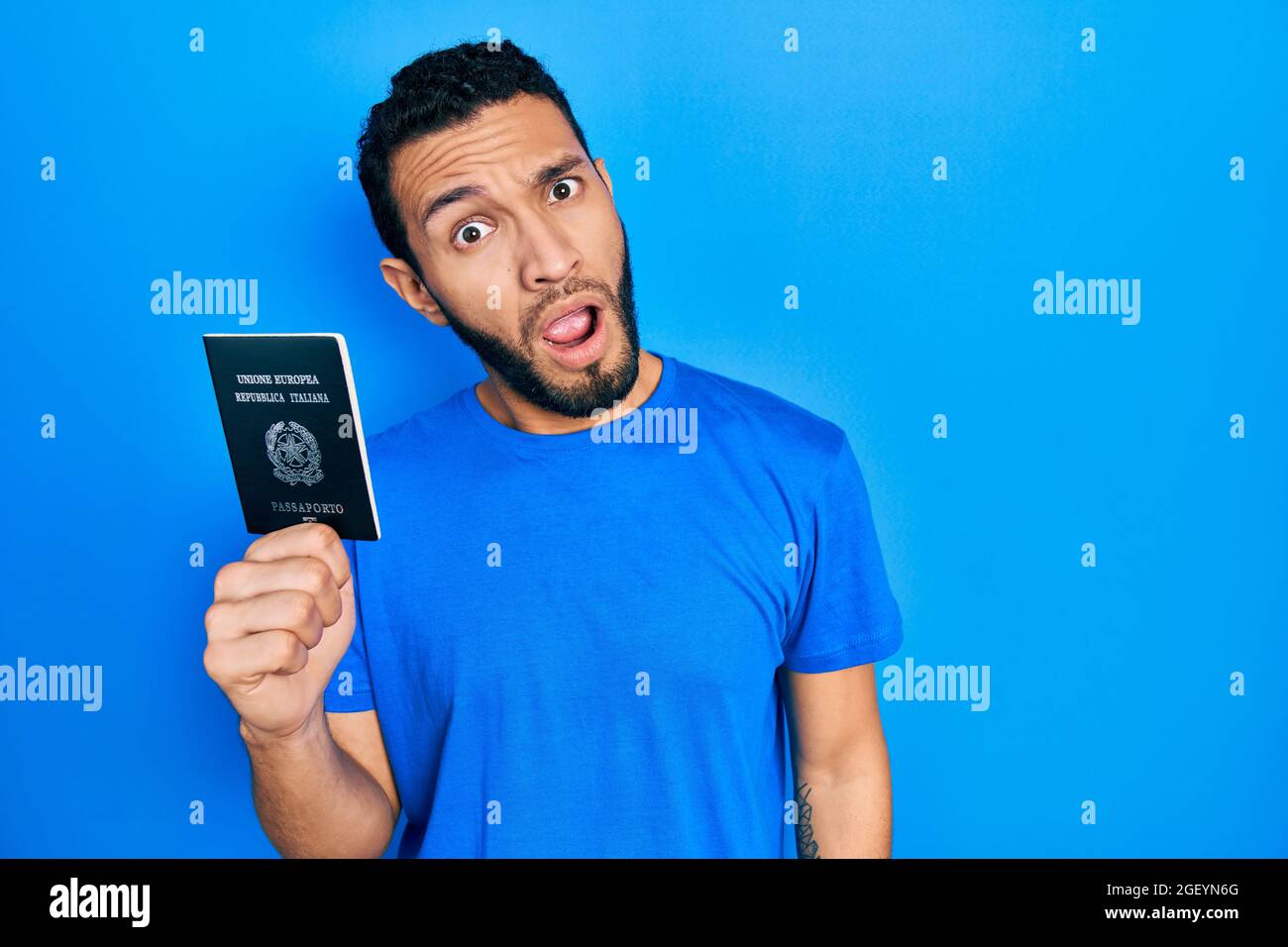 Uomo ispanico con barba che tiene il passaporto italia in faccia shock, che guarda scettico e sarcastico, sorpreso con bocca aperta Foto Stock