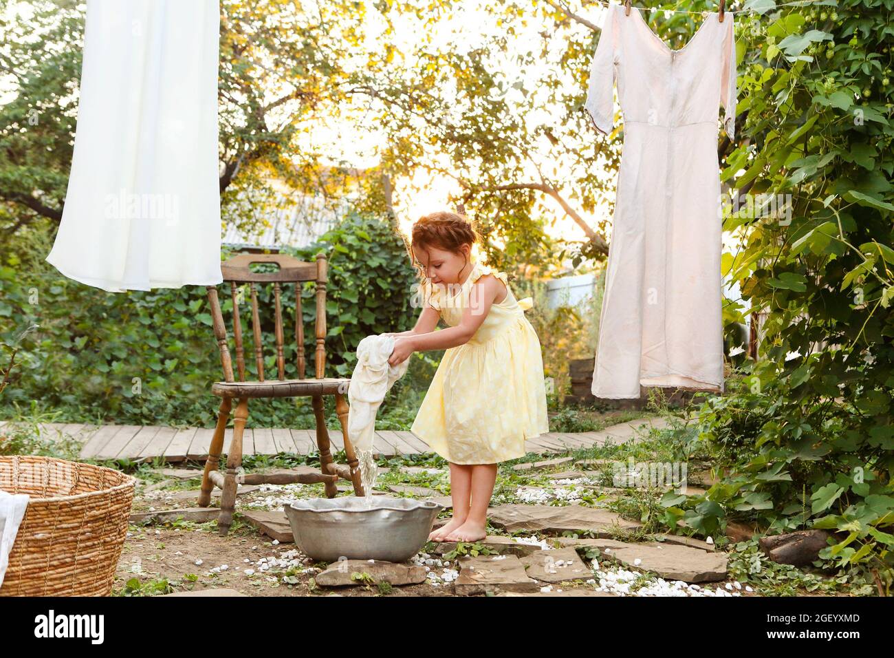 Ragazza piccola carina in vestito lavando i vestiti bianchi in un bacino di  metallo nel cortile,