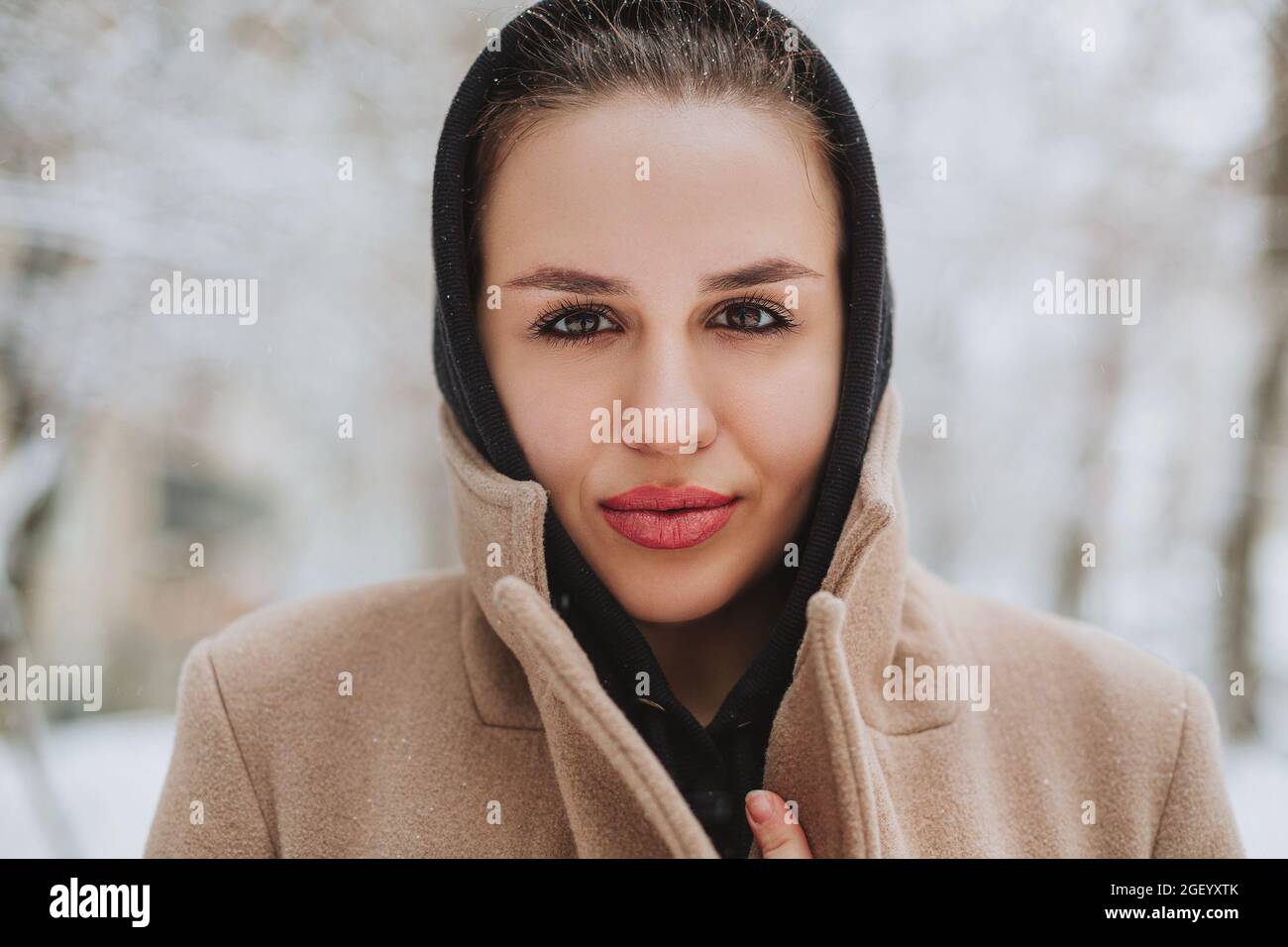 Giovane donna positiva con divertente sorriso sul viso si alza fuori in cappotto beige e sciarpa nera sulla testa, brivido dal freddo invernale contro bac Foto Stock