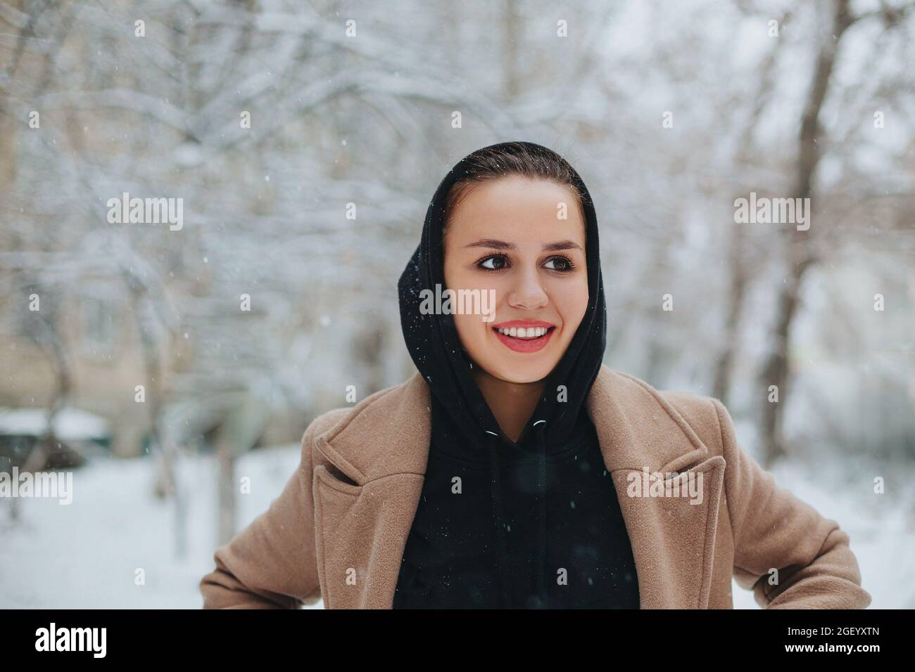 Giovane donna positiva con divertente sorriso sul viso si alza fuori in cappotto beige e sciarpa nera sulla testa, brivido dal freddo invernale contro bac Foto Stock