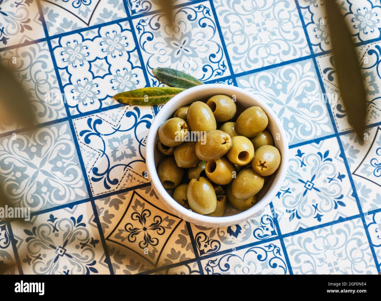 Piccola ciotola bianca con uno spuntino di olive verdi su un tavolo con piastrelle tradizionali blu mediterraneo sotto un ulivo Foto Stock