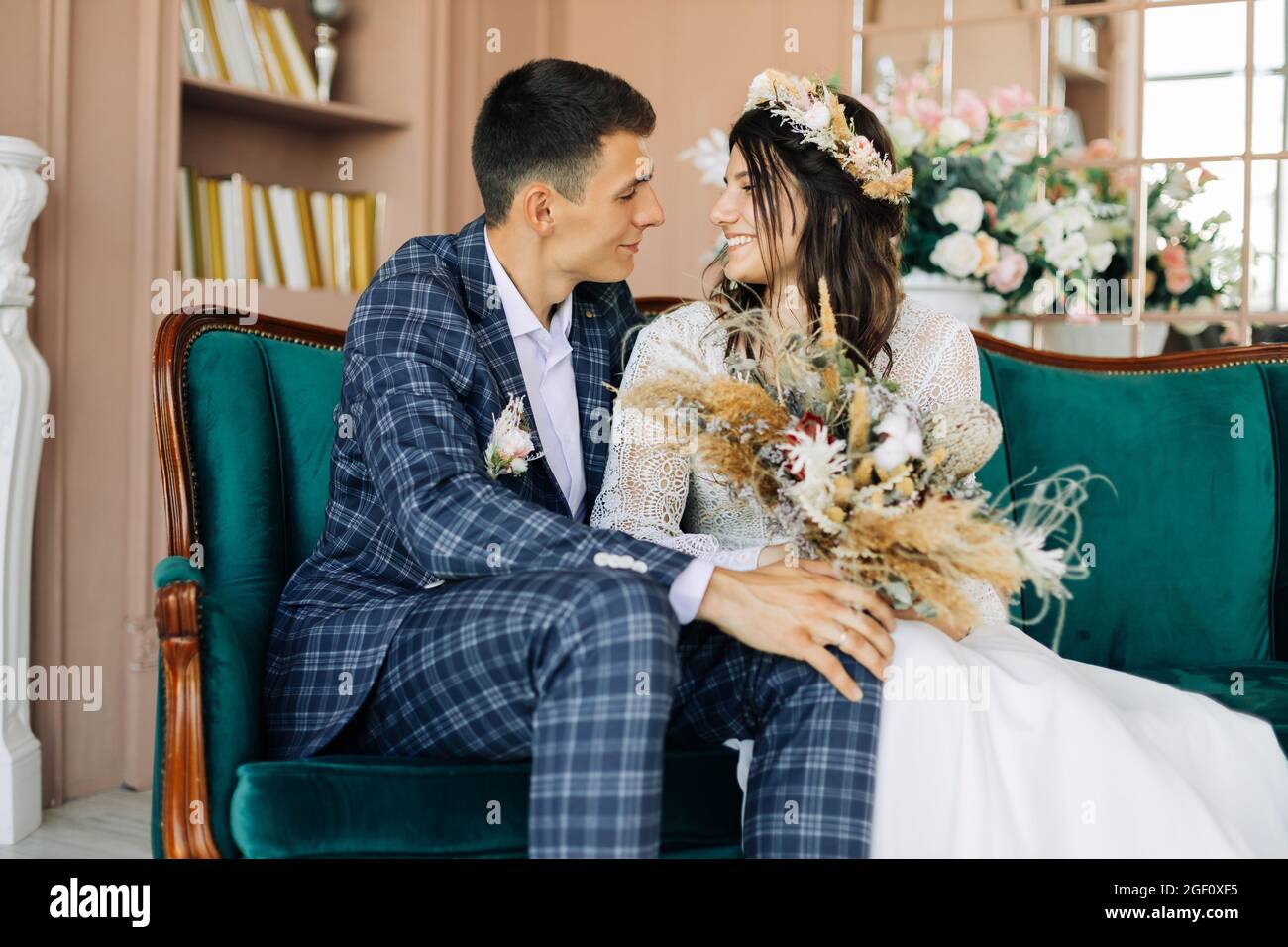 Felice coppia giovane, elegante sposo in un vestito e una giovane sposa in un abito da sposa con un bouquet di fiori, sedersi insieme, abbracciare, in studio su t Foto Stock