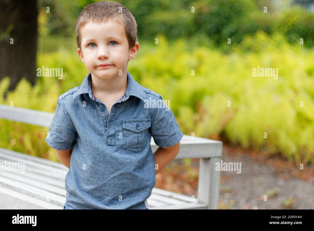 Primo piano di ragazzo carino in piedi accanto a una panchina in un parco verde in Polonia. Ritratto di un bel ragazzo biondo caucasico che guarda la fotocamera. Foto Stock