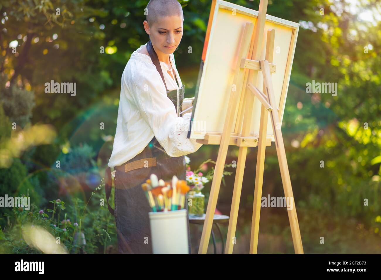 Bella giovane donna che si rilassa mentre dipinge una tela d'arte all'aperto nel suo giardino. Superstite da cancro, consapevolezza, arte terapia, concetto di creatività. Foto Stock