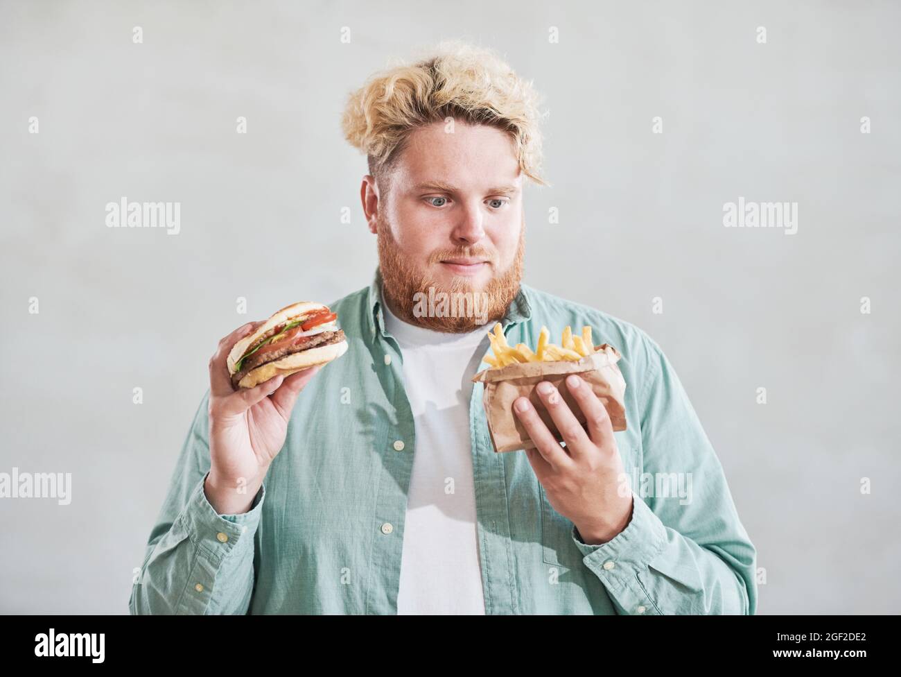 Giovane uomo sovrappeso che tiene patatine fritte in una mano e hamburger in un'altra mano isolato su sfondo bianco Foto Stock