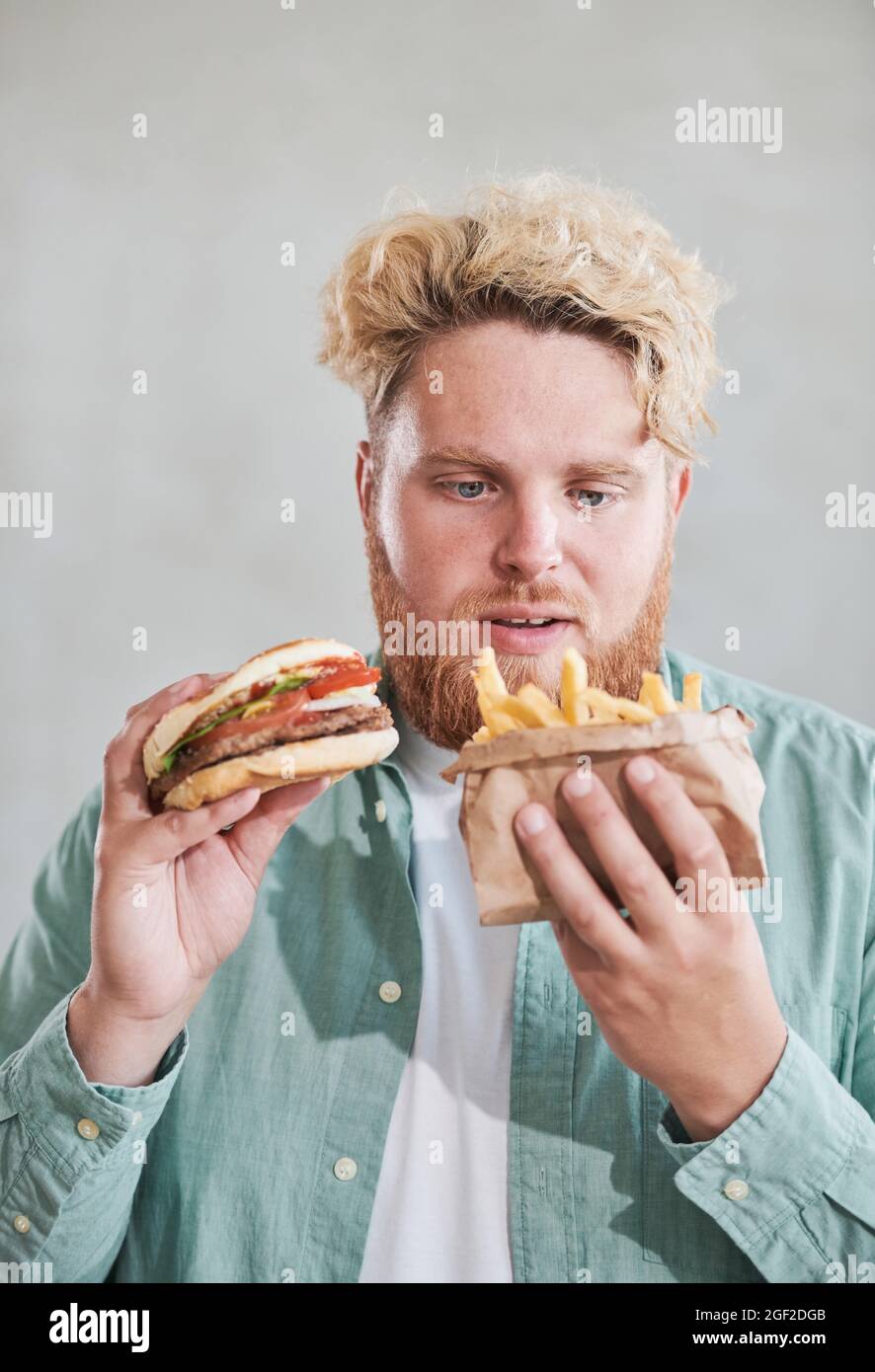 Uomo sovrappeso che mangia patatine fritte e hamburger su sfondo bianco Foto Stock