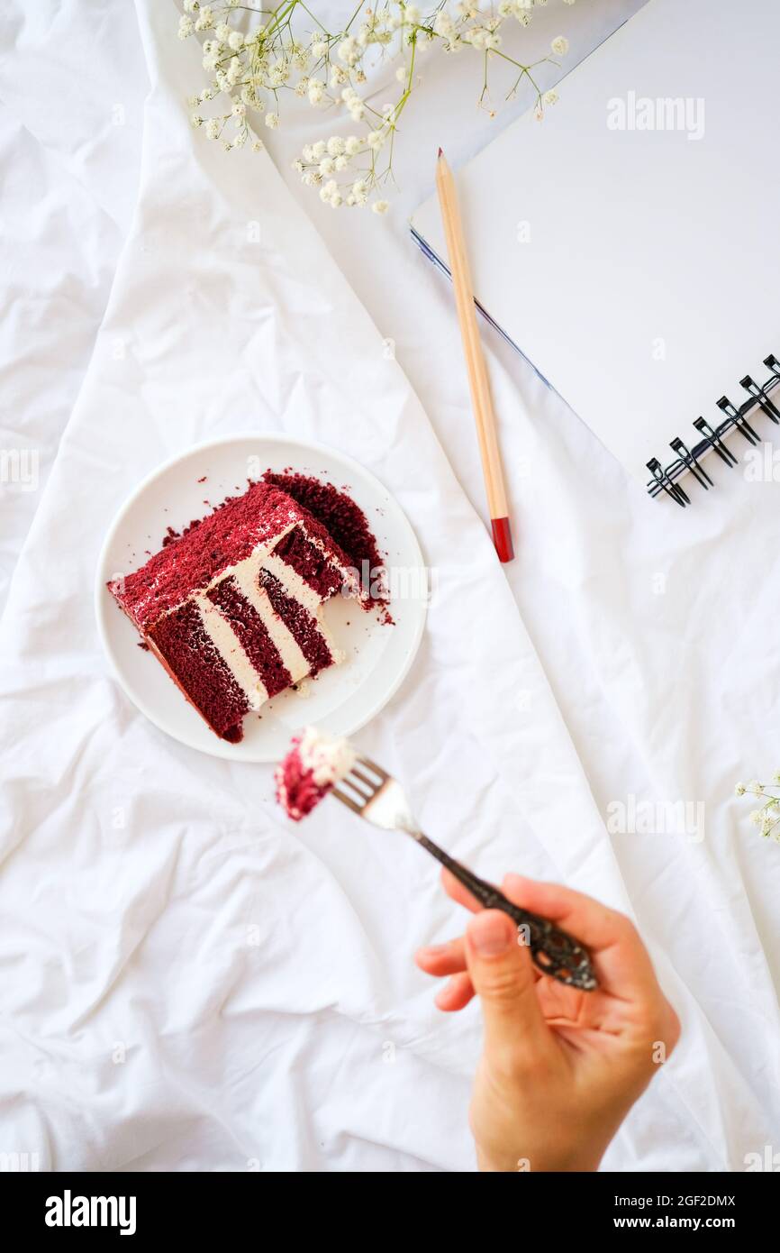 Flat Lay donna mani che tengono forchetta per mangiare pezzo di torta di  velluto rosso su fogli bianchi a letto, taccuino, matita e fiore intorno.  Concetto di stile di vita estetico Foto