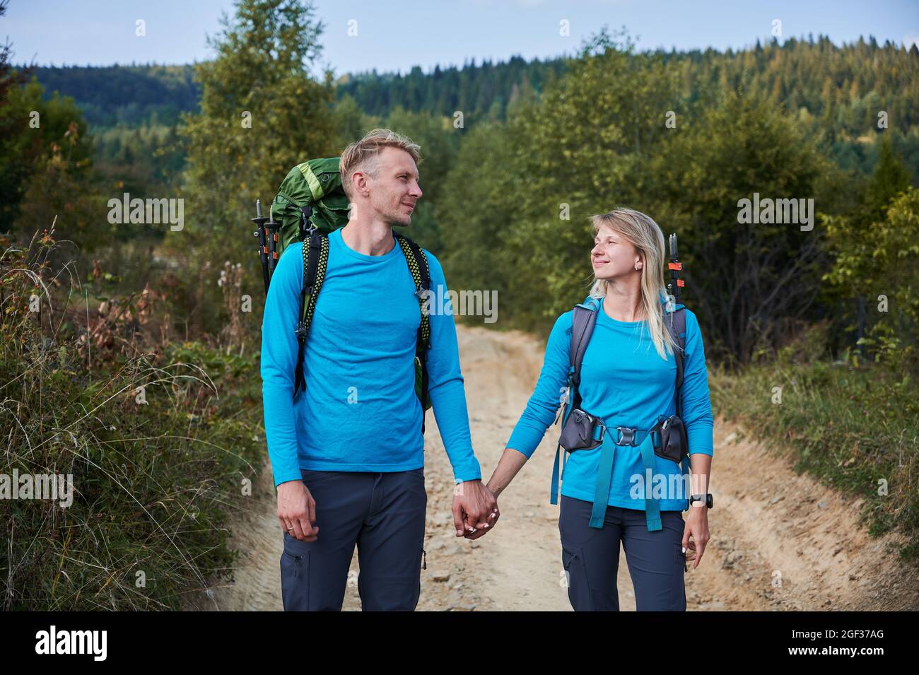 Coppia amante di turisti negli stessi abiti con zaini che camminano su strada sterrata all'aperto, tenendo le mani l'un l'altro. Due escursionisti camminano lentamente sul sentiero sullo sfondo di erba verde e foreste. Foto Stock