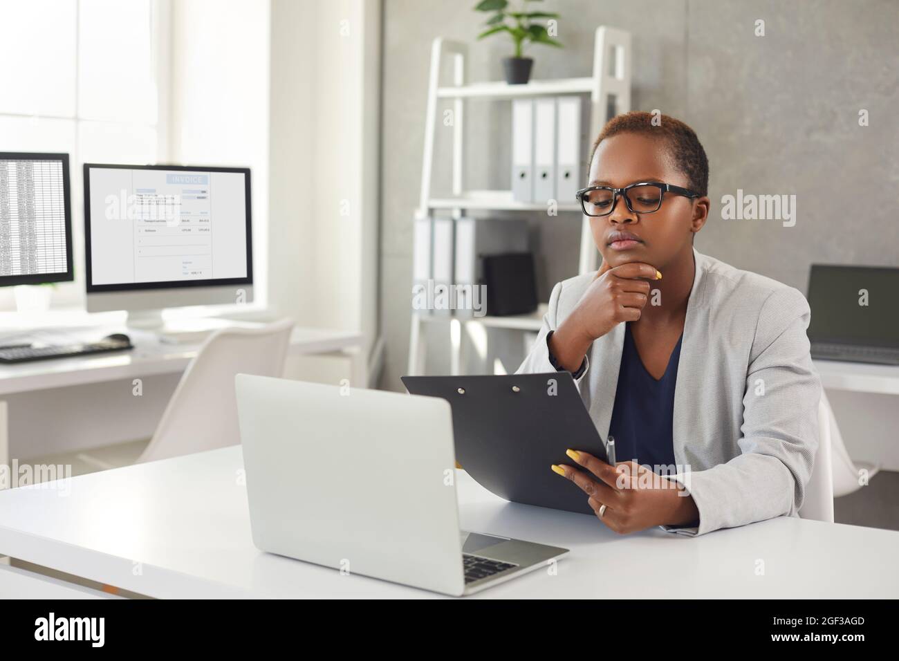 Donna nera seduta alla sua scrivania in ufficio, lettura di documenti aziendali e pensiero Foto Stock