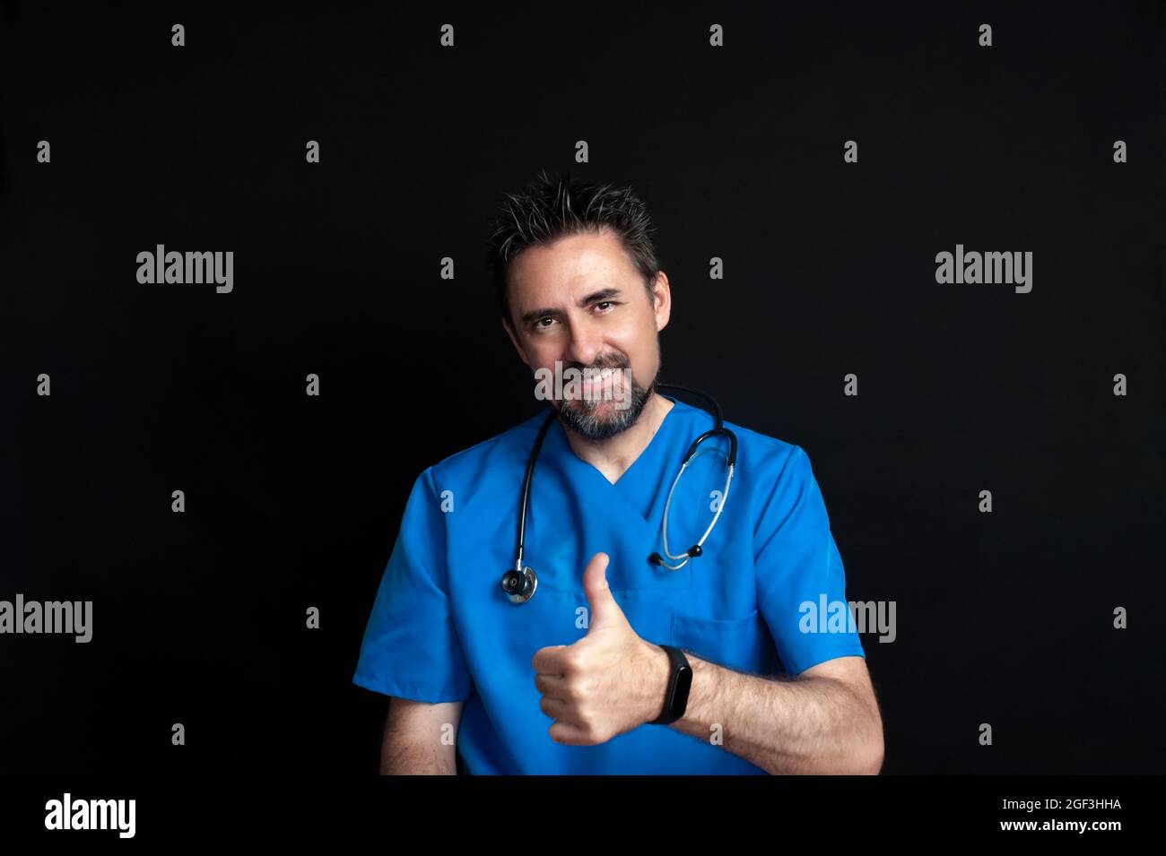 Un medico maschio vestito in una uniforme blu dell'ospedale, scuro e barbuto, portando uno stetoscopio intorno al collo, sorridendo e dando un pollice su indicando t Foto Stock