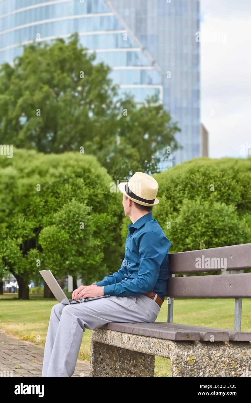 Un uomo con un computer portatile si siede su una panchina in un parco cittadino, lavora a distanza e guarda verso un edificio di uffici nel centro della città. Foto Stock