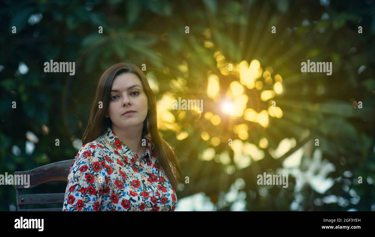 Bella giovane donna latina in abito floreale di design seduto in giardino guardando direttamente nella macchina fotografica con raggi del sole al tramonto che passa attraverso Foto Stock