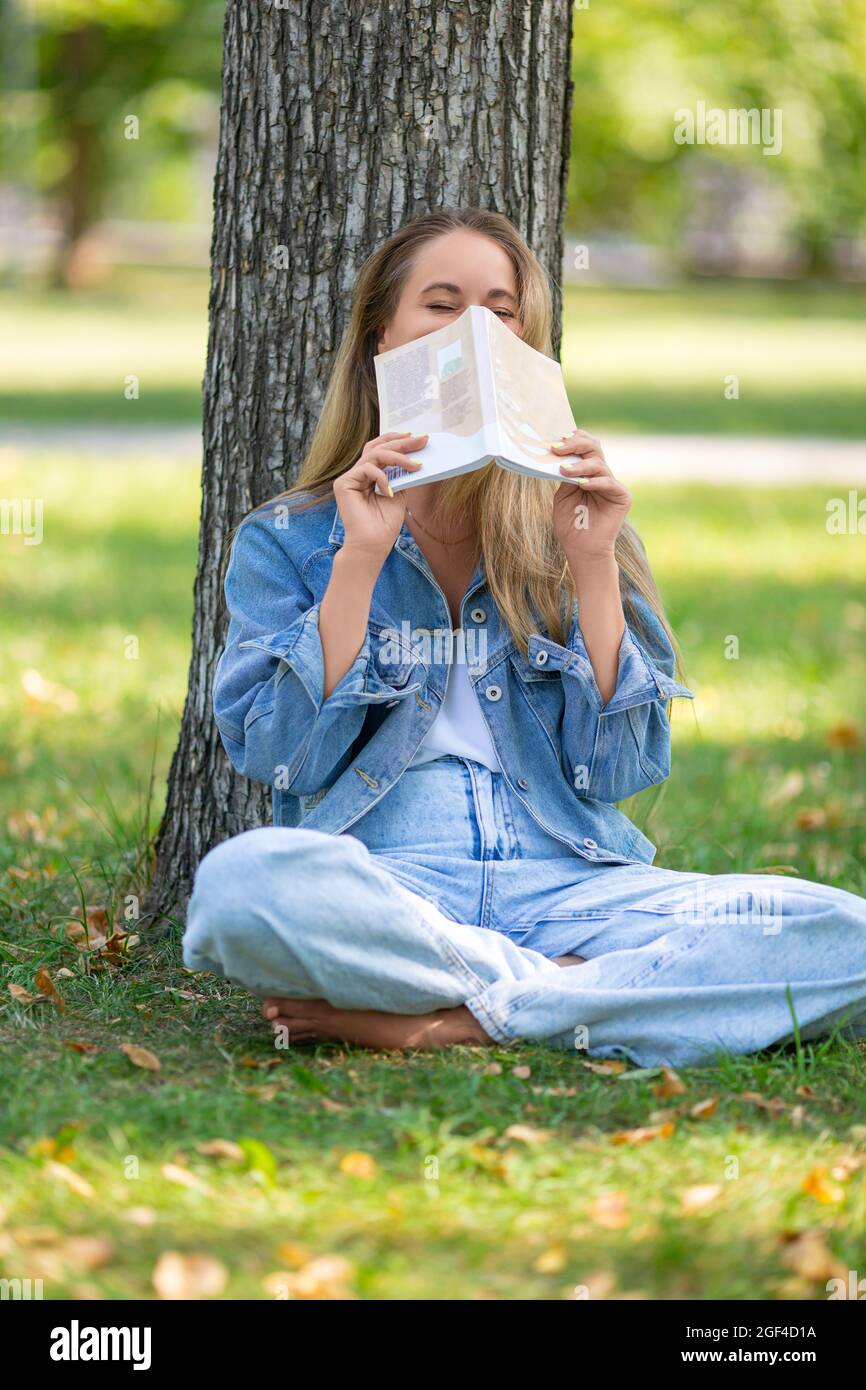 Foto verticale di una donna con un libro nel parco. Lei si siede sull'erba e ride, coprendo il viso con un libro. Concetto di riposo da gadget e c Foto Stock