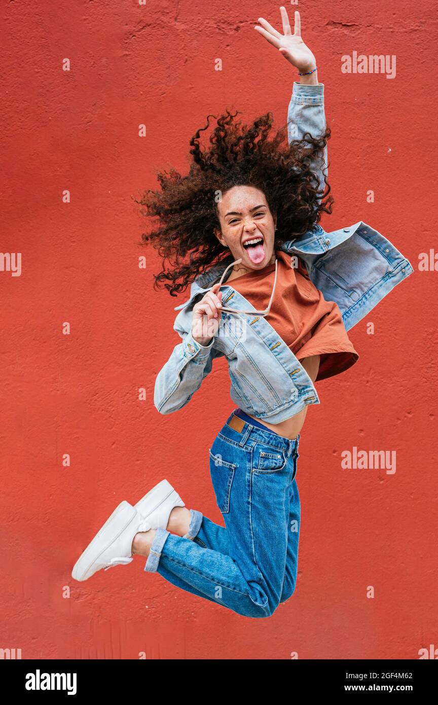Giovane donna allegra che attacca la lingua mentre salta di fronte al muro rosso Foto Stock