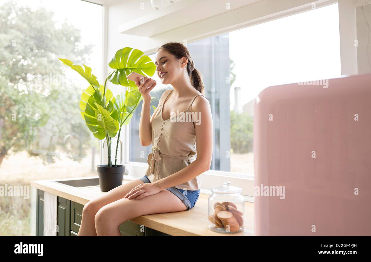 Giovane donna che invia messaggi vocali tramite smartphone mentre si siede sul bancone della cucina a casa Foto Stock
