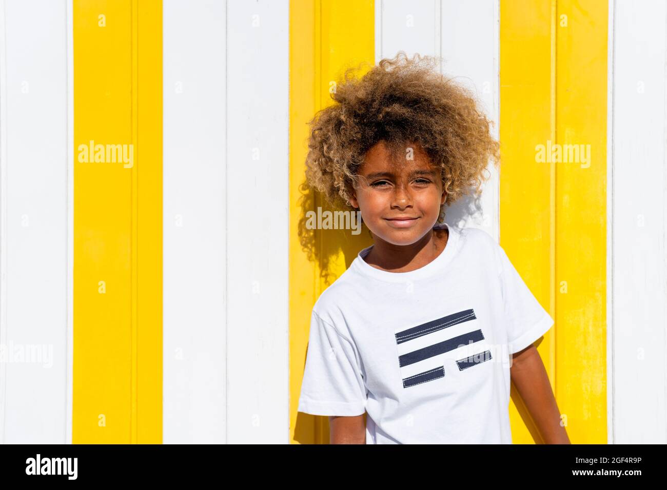 Ragazzo sorridente con capelli ricci davanti a una parete bianca e gialla a righe Foto Stock