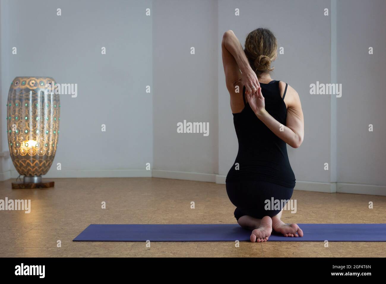 Indietro yoga femminile seduta in gomukhasana su tappeto con lampada sul retro. Fit donna pratica yin yoga faccia posa in studio. Stile di vita sano, abbigliamento attivo Foto Stock