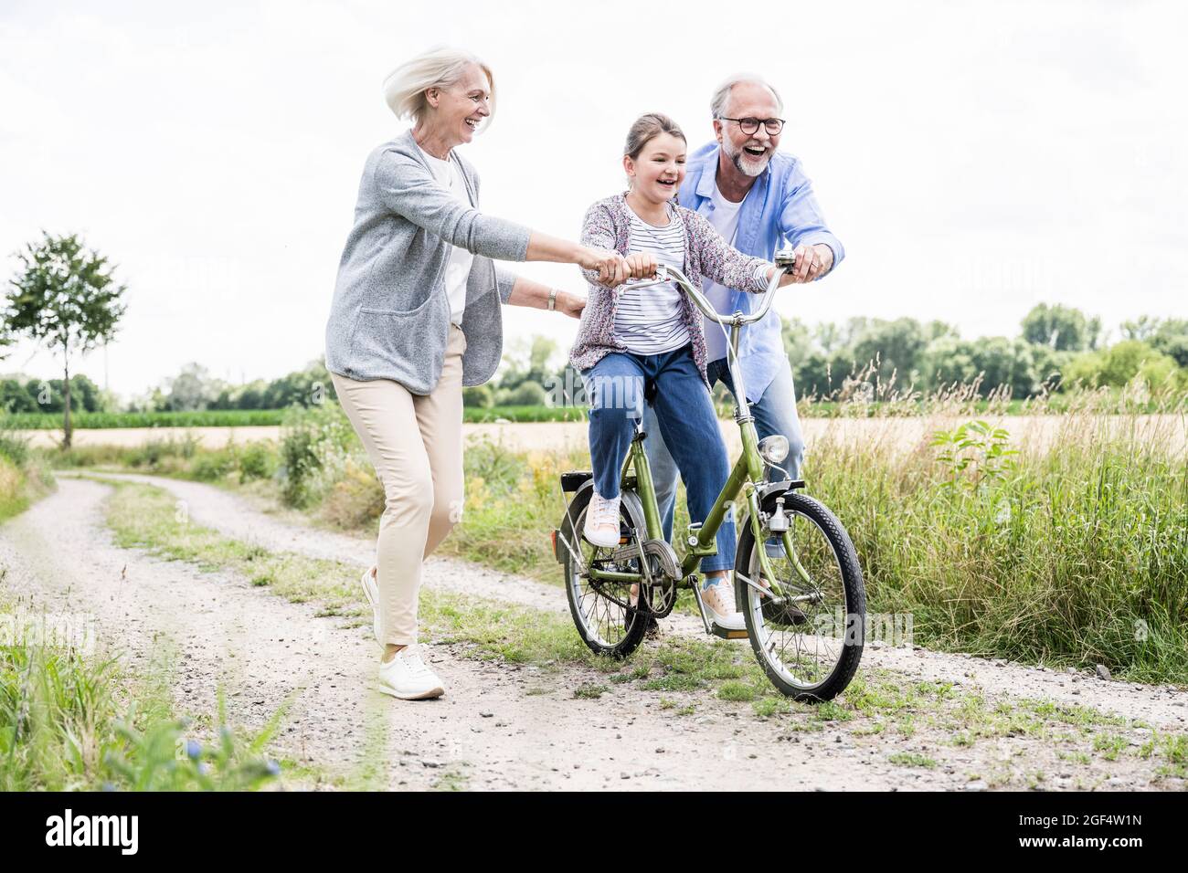 Nonno allegro aiutando nonna mentre si guida in bicicletta su strada sterrata Foto Stock