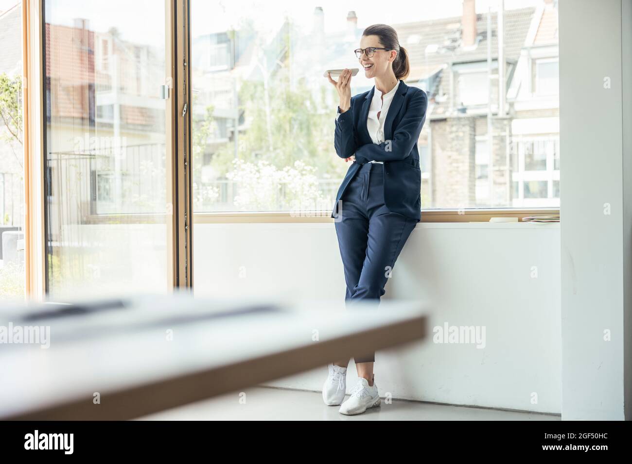 Donna d'affari che invia messaggi vocali tramite smartphone mentre si appoggia alla finestra Foto Stock