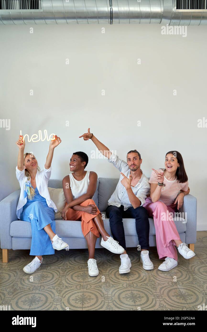 Donna d'affari che tiene Ciao segno di parola mentre si siede con i colleghi sul divano Foto Stock