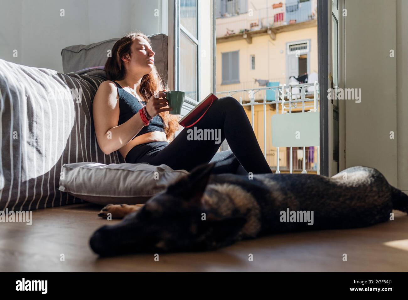 Donna con tazza di caffè e libro rilassante da cane a casa Foto Stock