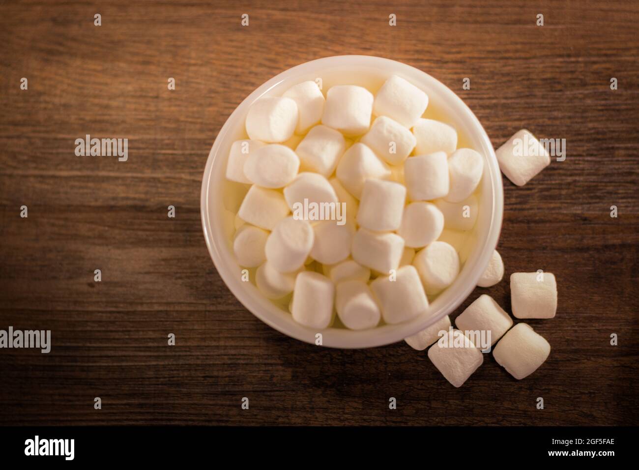 Un primo colpo di una ciotola di piccoli marshmallow bianchi con marshmallow caduto sul tavolo di legno, scatto dello studio della ciotola di marshmallow sul tavolo della cucina Foto Stock