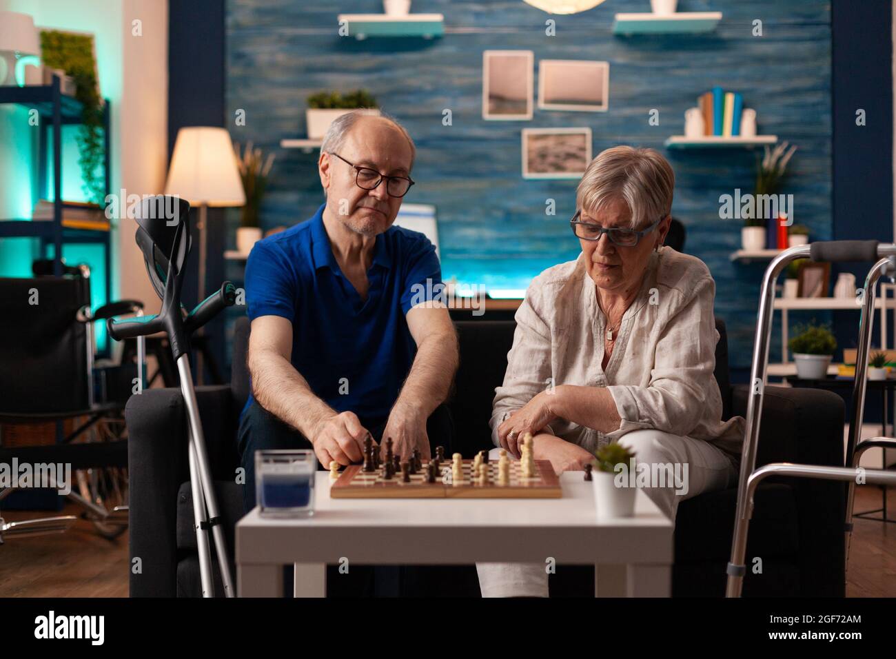 Coppia anziana che gioca a scacchi a bordo seduti a casa insieme sul divano. Vecchio uomo e donna in pensione maturo con crutches Walk Frame e sedia a rotelle godendo il gioco di mente per l'intrattenimento Foto Stock