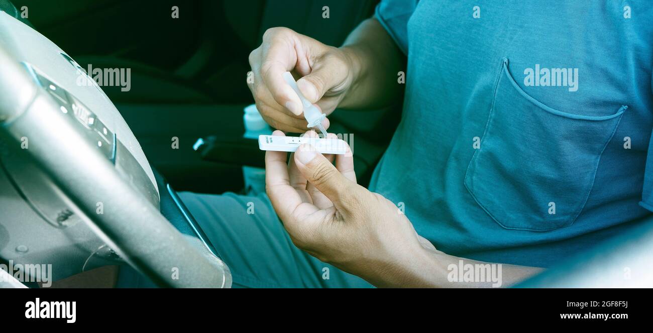 un giovane uomo pone il proprio campione nel dispositivo di test diagnostico dell'antigene covid-19 mentre è seduto sul sedile del conducente della sua auto, in una forma panoramica Foto Stock