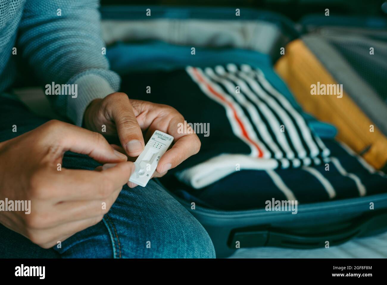 un giovane uomo, indossando abiti casual, colloca il proprio campione nel dispositivo di test diagnostico dell'antigene covid-19 seduto sul letto accanto alla sua valigia, confezione Foto Stock