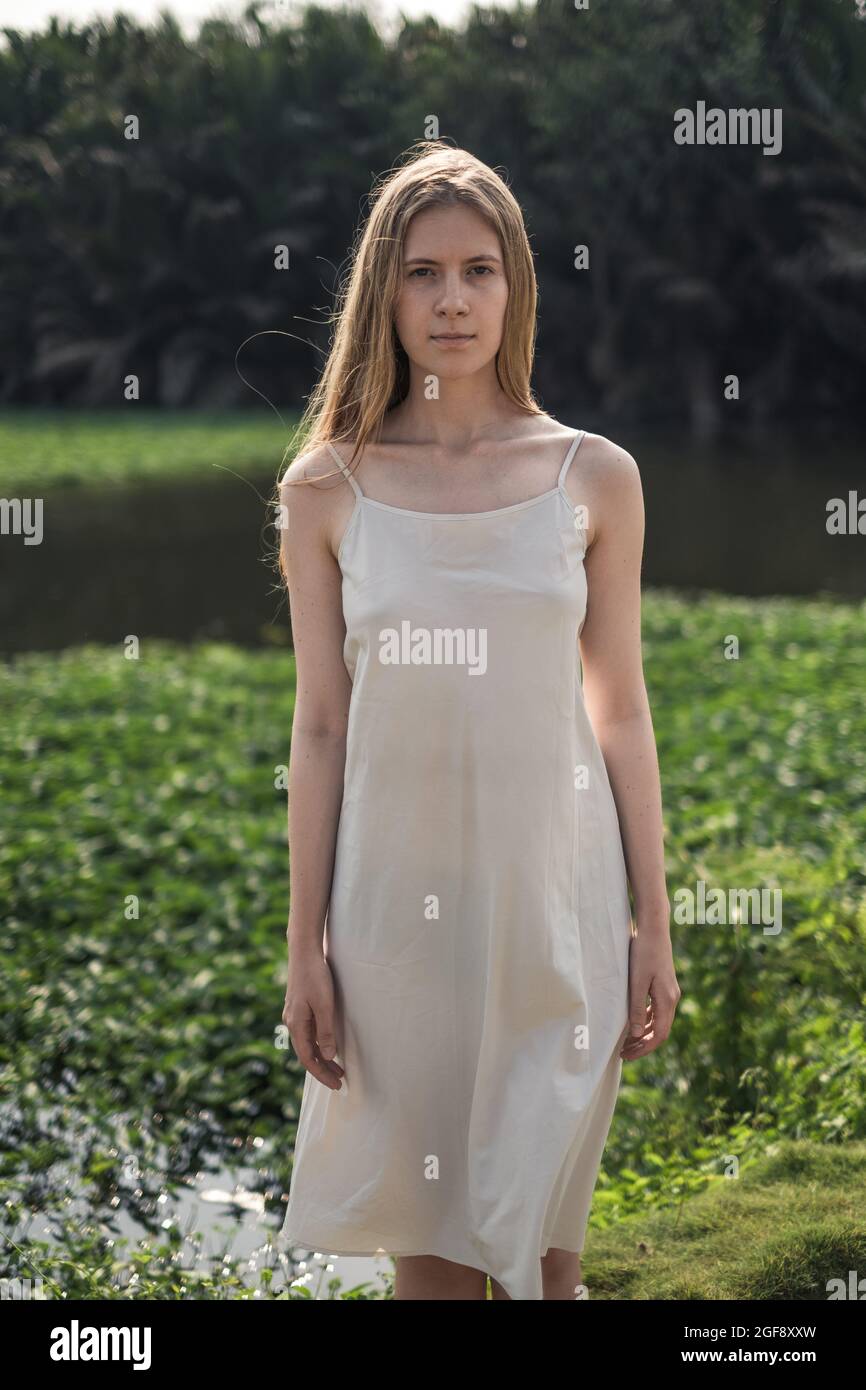Ritratto di attraente giovane donna caucasica in abito bianco in piedi nel parco. Capelli biondi lunghi. Bellezza naturale. Il make-up è gratuito. Foto Stock
