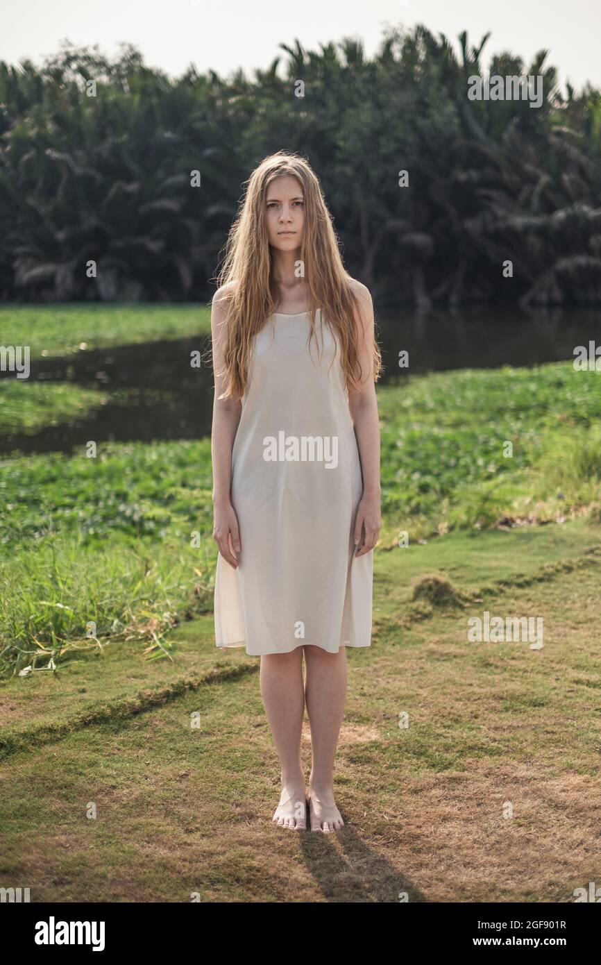 Ritratto a tutta lunghezza di giovane graziosa donna caucasica in abito bianco in piedi a piedi nudi nel parco. Capelli biondi lunghi. Bellezza naturale. Il make-up è gratuito. Foto Stock