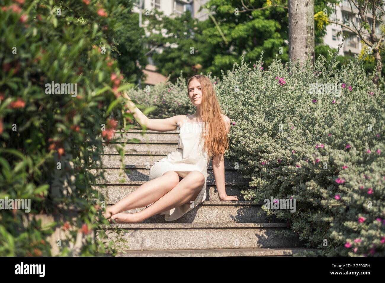 Bella giovane donna caucasica in abito bianco seduto sulle scale nel parco. Capelli biondi lunghi. Belle piante. Bellezza naturale senza make-up Foto Stock