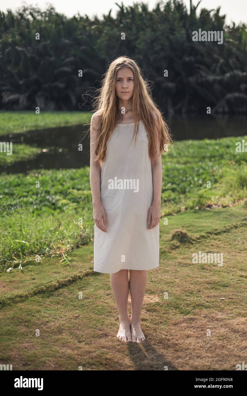 Giovane bella donna caucasica in abito bianco in piedi nel parco. Capelli biondi lunghi. Bellezza naturale. Il make-up è gratuito. Bellissimo sole Foto Stock