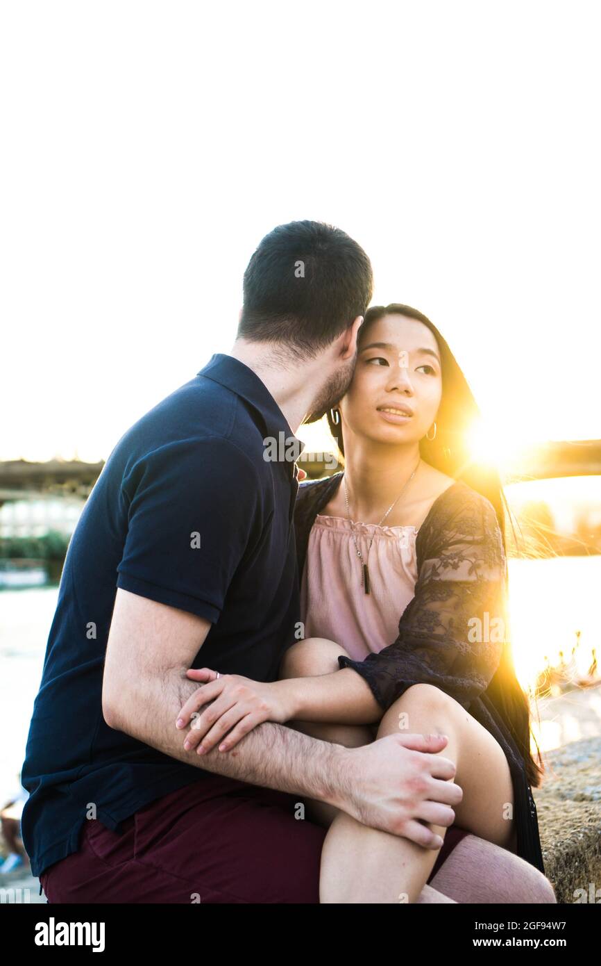 Giovane coppia multirazziale seduta sul fiume con un tramonto sullo sfondo. Uomo che bacia la sua ragazza in una giornata estiva. Ragazza cinese con lei Foto Stock