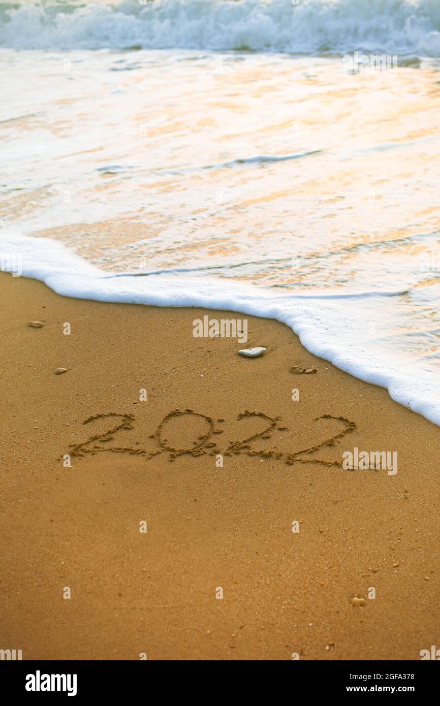 2022 Happy New Year iscrizione spiaggia di sabbia. Il testo scritto è scritto su sabbia gialla ed è lavato via da un'onda. Il concetto di cambiamento dell'anno, Foto Stock