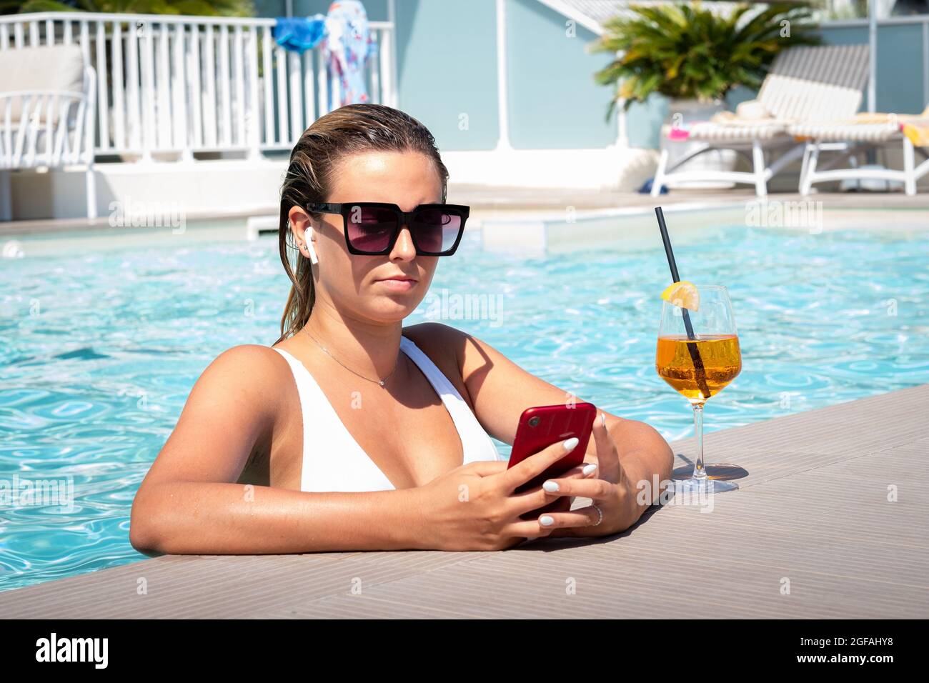 Ragazza adolescente videocalling con uno smart phone rosso mentre si fa il bagno in piscina in vacanze estive - Donna al telefono SMS mentre si rilassa in po Foto Stock