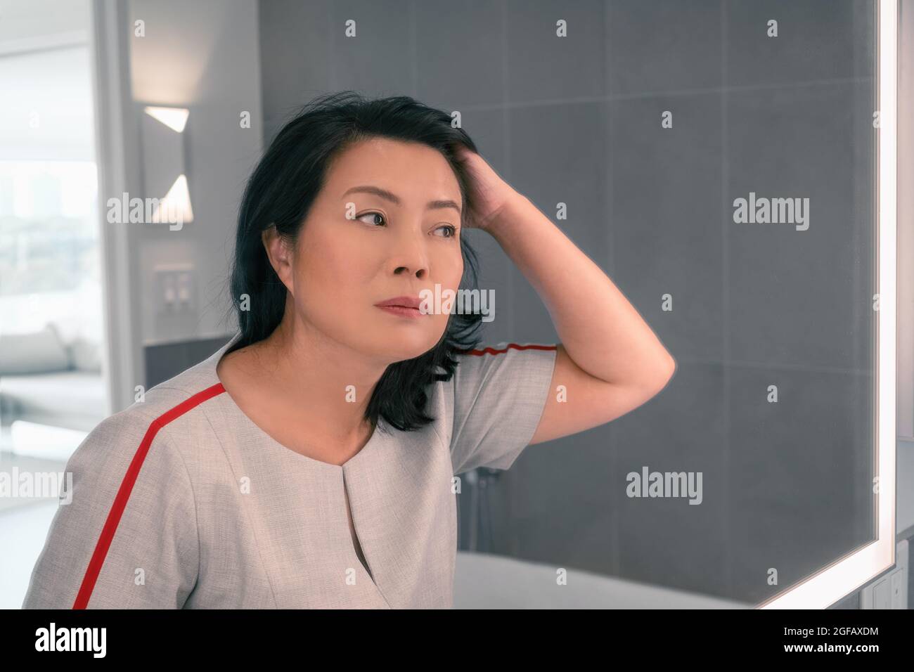Perdita di capelli matura donna asiatica che tocca i suoi capelli styling o colorazione capelli grigi guardando se stessa nello specchio di casa. Beautifiul signora professionale cinese Foto Stock