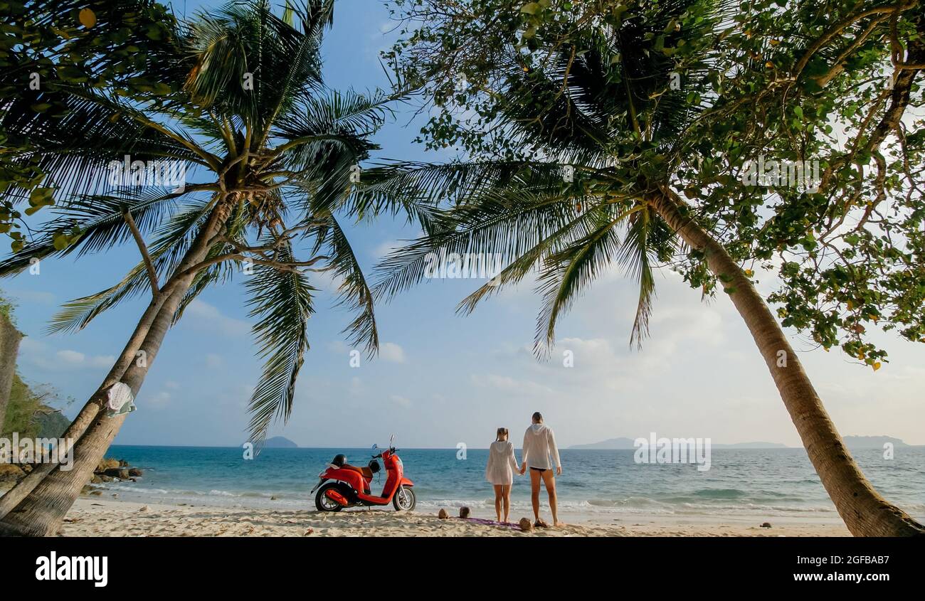 Viaggio su strada in scooter. Bella coppia su moto rossa in abiti bianchi sulla spiaggia di sabbia. Persone che camminano vicino alle palme tropicali, mare. Noleggio moto. Foto Stock