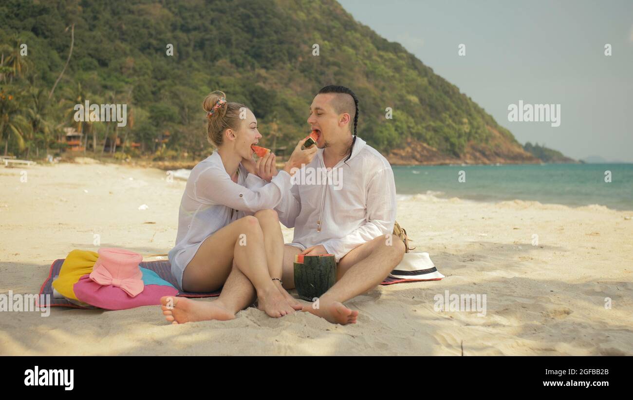 La allegra coppia d'amore che tiene e mangia fette di anguria sulla spiaggia tropicale di sabbia del mare. Amanti romantici due persone trascorrono il fine settimana estivo. Foto Stock