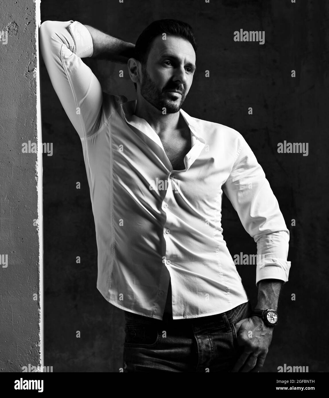 Bianco e nero. Uomo adulto non rasato in camicia bianca e jeans si erge su una parete di cemento, appoggiandosi, tenendo la mano dietro la testa Foto Stock