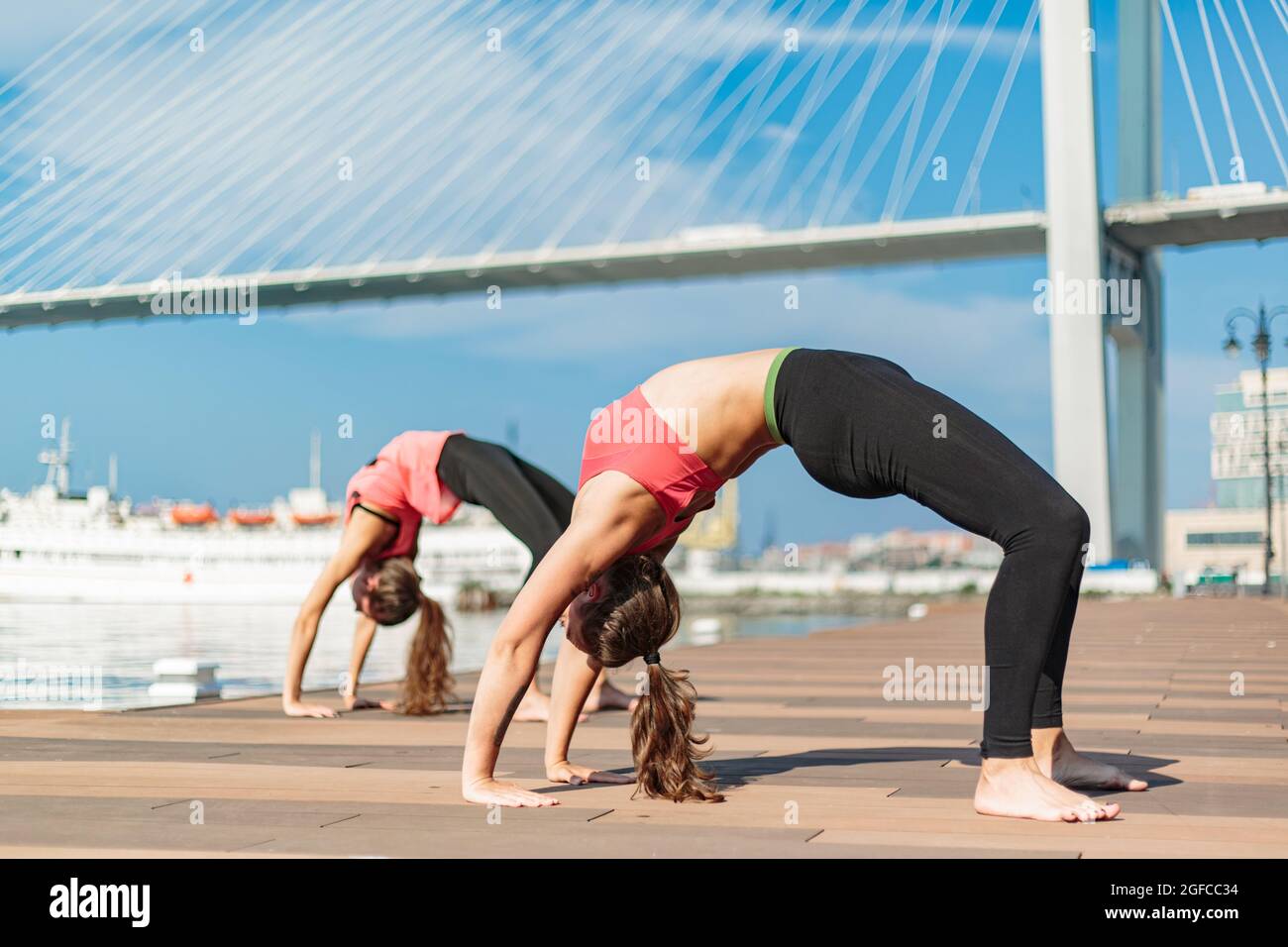 Due ragazze giovani in tute identiche fanno un elemento di yoga. Stanno facendo yoga nello sfondo della città. È una giornata di sole. Yoga sul molo n Foto Stock