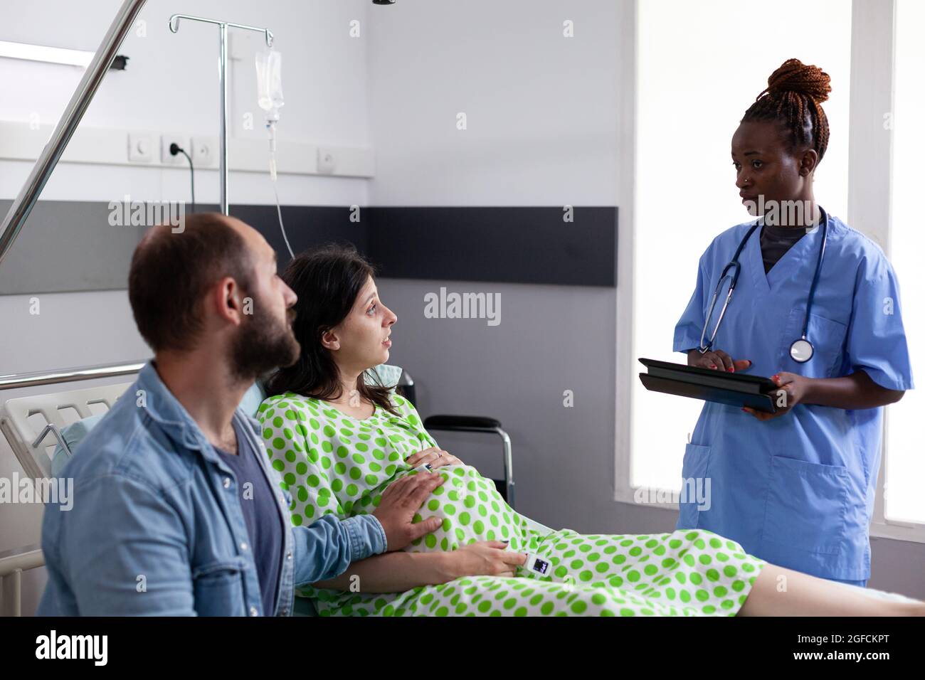 La coppia caucasica si aspetta un bambino in ospedale mentre parla con l'infermiera afroamericana alla maternità. Genitori con gravidanza che ricevono assistenza medica preparazione per la consegna del bambino Foto Stock
